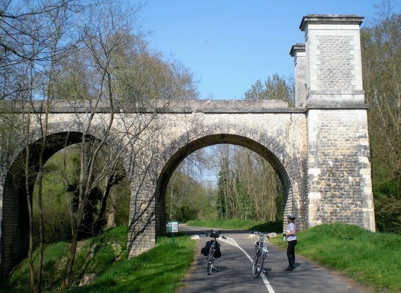 woman standing next to two bicycles near a scenic bridge in france