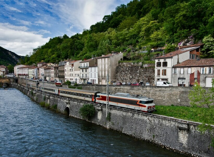 train riding along between a river, a city street with mountains in the background near Foix