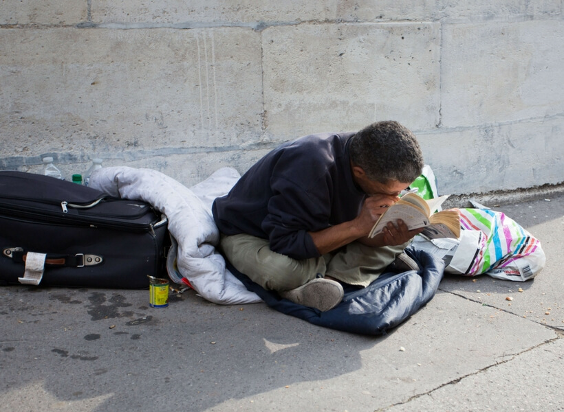 beggar sitting on a paris street surrounded by his few posessions, he is observing a book at close distance, possibly a visually impaired person