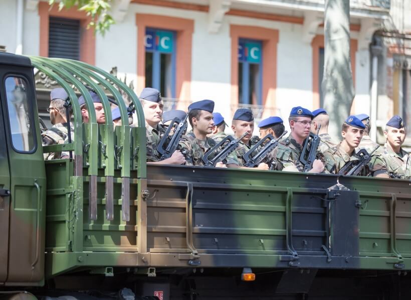 military personnel sitting on the bed of a truck with their machine guns across their chests