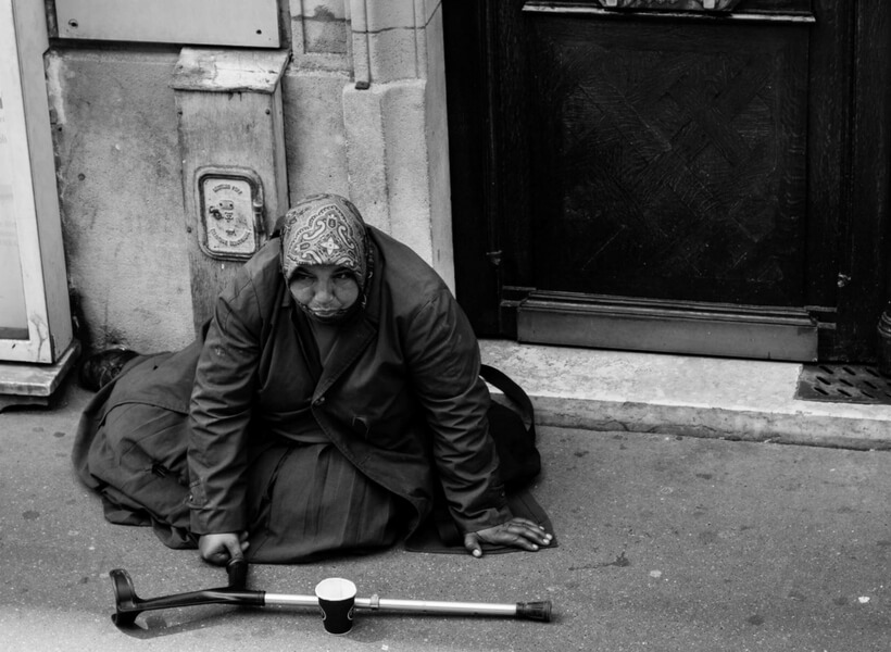 woman begging on the streets of paris, she's wearing a heavy coat and a head covering and has a crutch and paper cup in front of her for donations